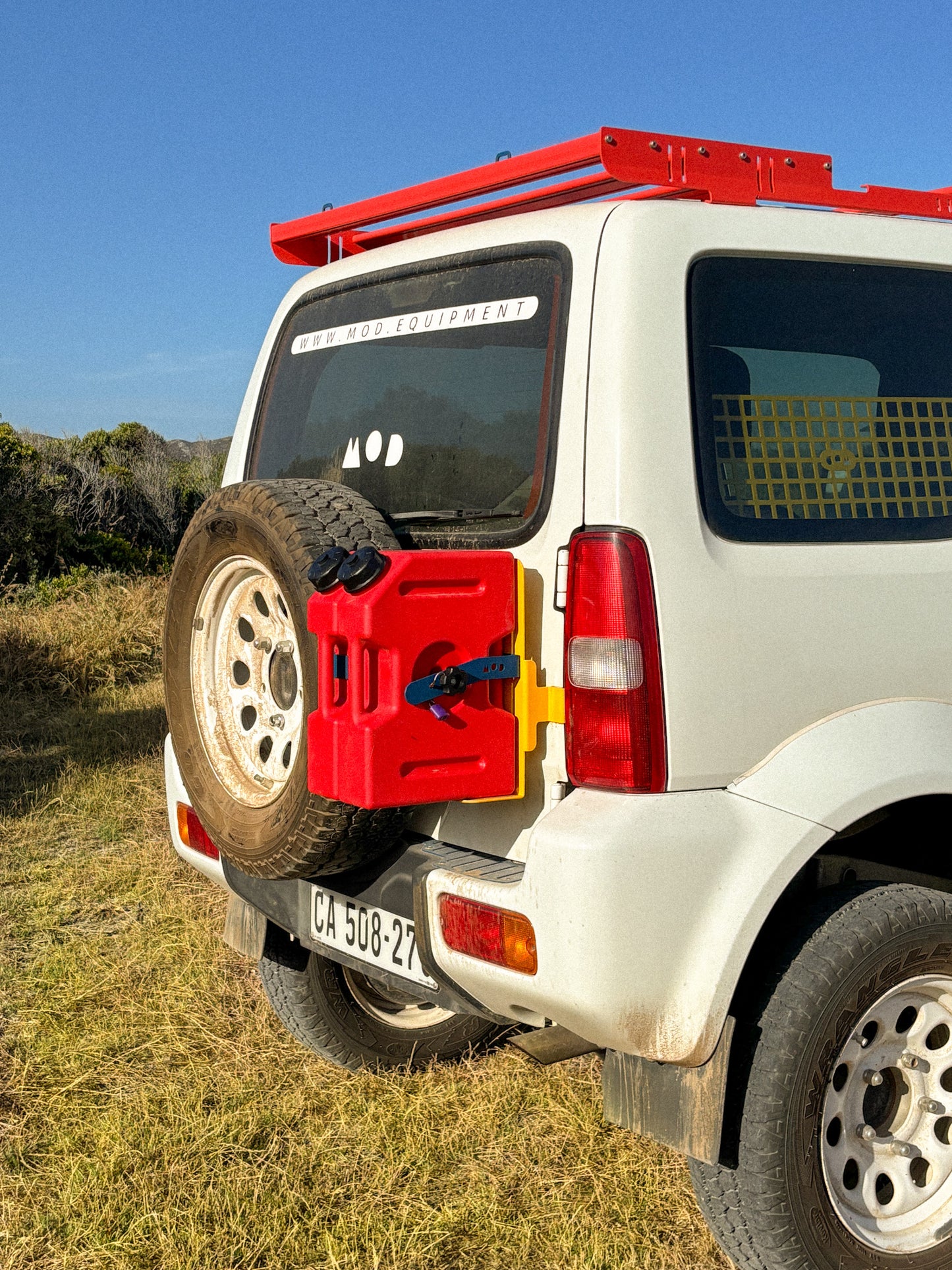 White Suzuki Jimny 3 with a red fuel tanks mounted on the rear door on a grassy field