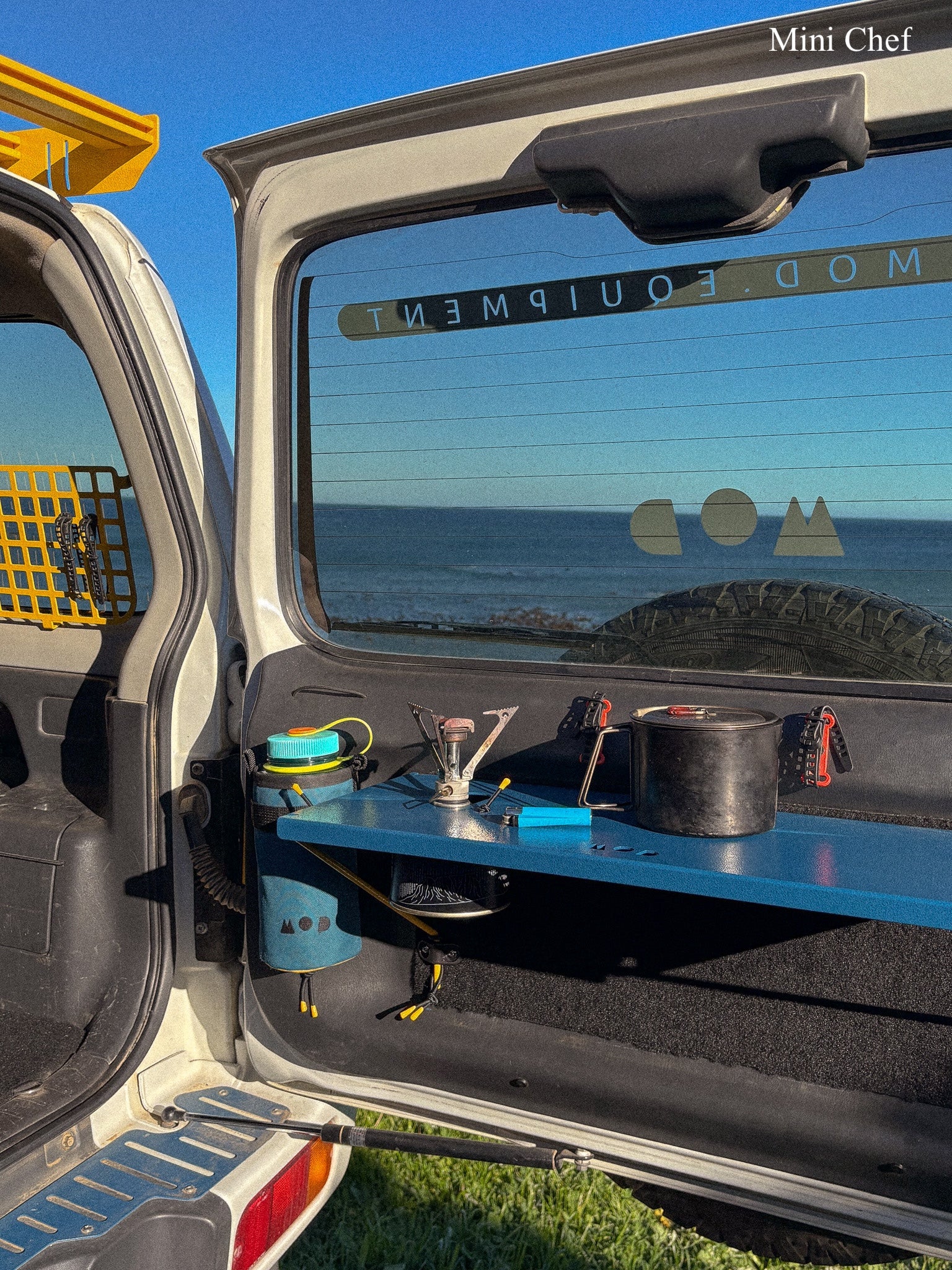 An outdoor setup inside the open rear door of a vehicle, with a blue rear door table holding camping gear including a black pot, stove, and accessories. The vehicle window shows the ocean and sky behind it.