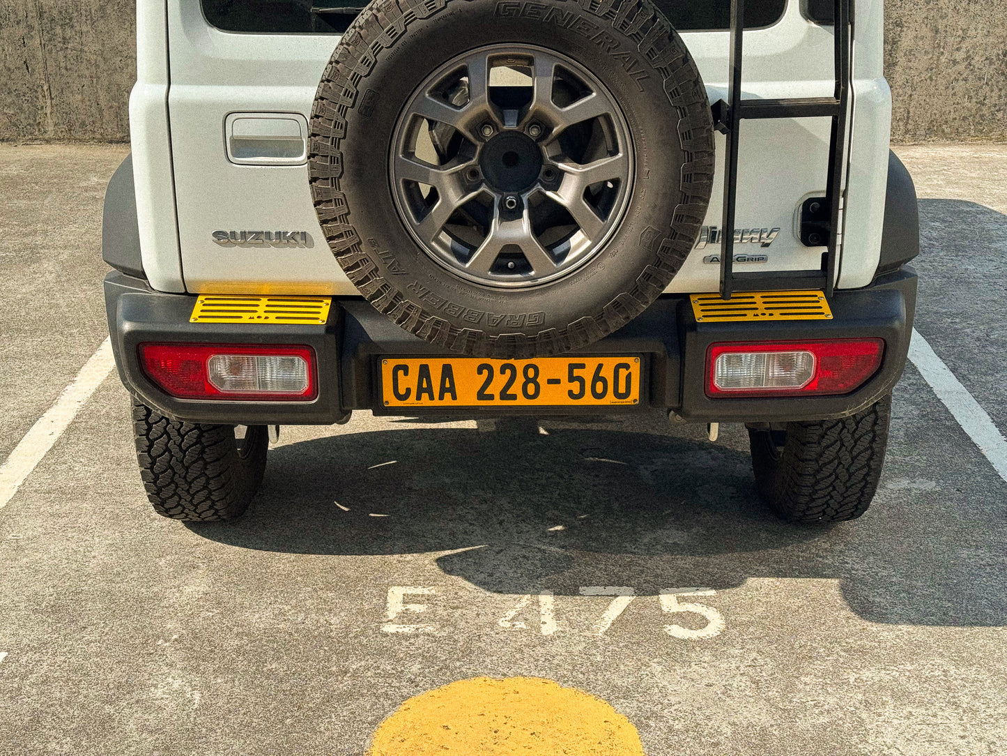 Rear view of a white Suzuki Jimny 4 parked on a concrete surface, showing the spare tire mounted on the back, rear bumper with yellow MOD tread plates installed