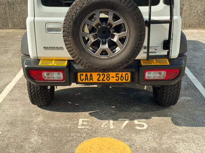 Rear view of a white Suzuki Jimny 4 parked on a concrete surface, showing the spare tire mounted on the back, rear bumper with yellow MOD tread plates installed