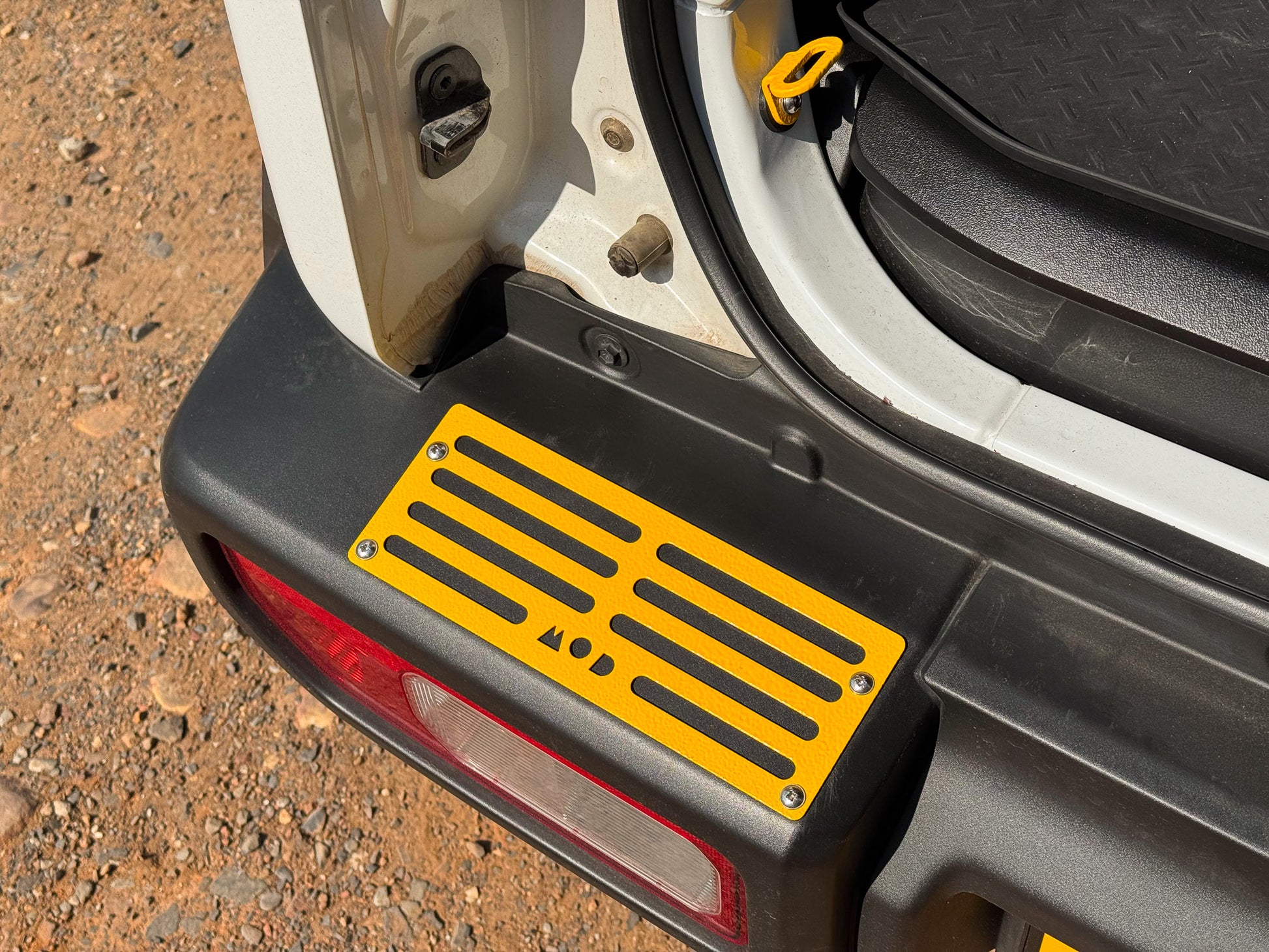 A close-up of a bright yellow MOD tread plate installed on a black Jimny rear bumper, with the open car door and gravel ground visible.