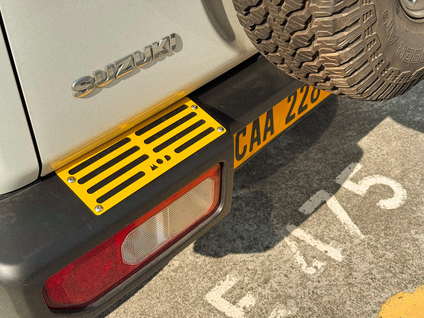 Close-up of the rear left side of a Suzuki Jimny 4, showing a bright yellow MOD tread plate mounted on the bumper